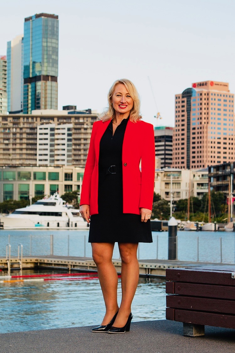 Genevieve Sage in an orange jacket, standing by the waterfront with city buildings in the background